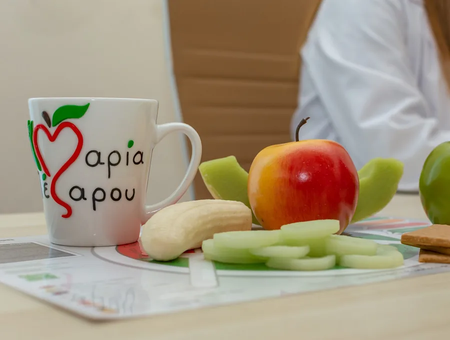 Κούπα με το λογότυπο της Μαρίας Πέζαρου, δίπλα σε φρέσκα φρούτα